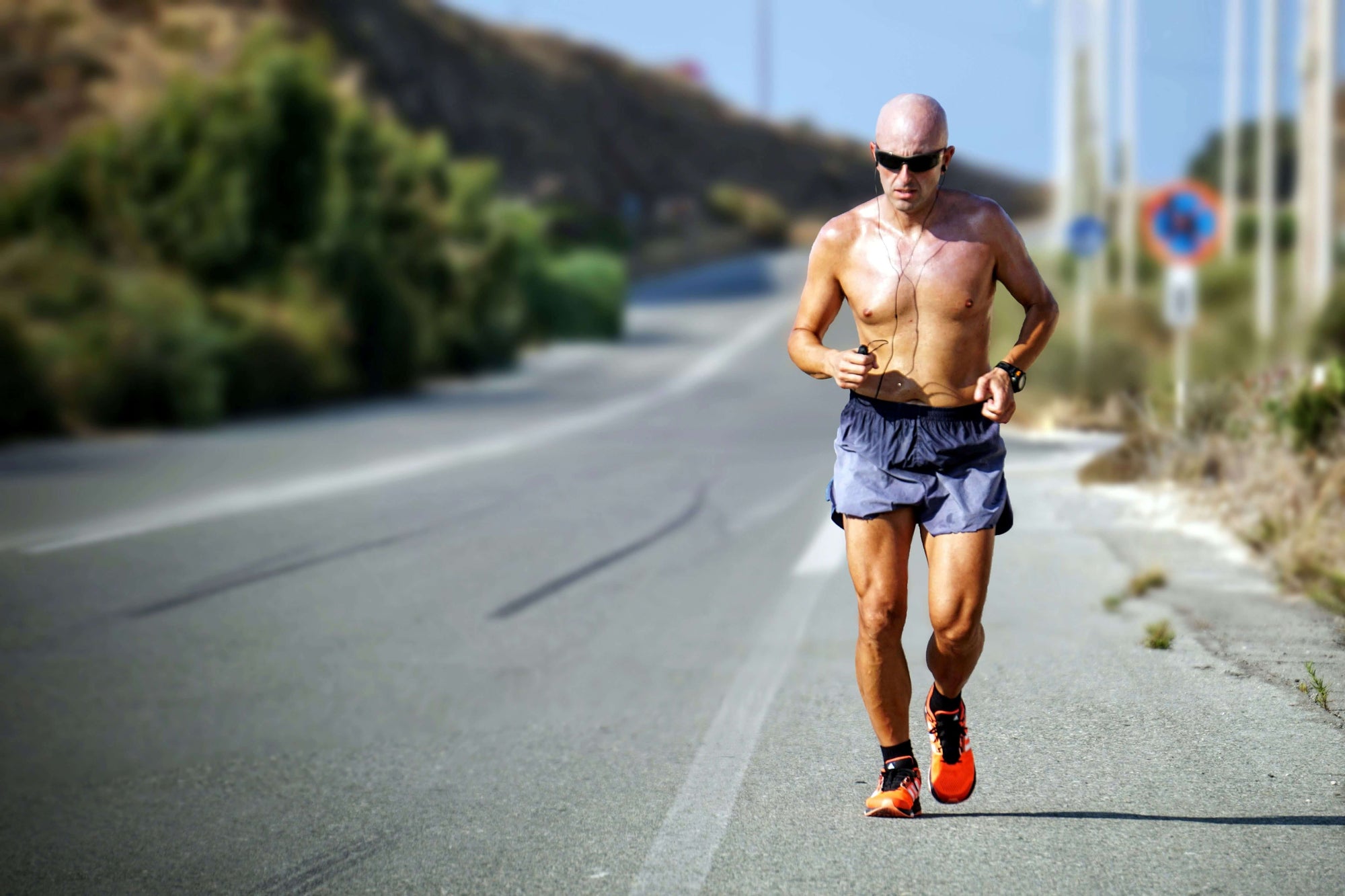 Shirtless man wearing blue shorts as one of the types of running pants, running on a road and wearing black earphones. 