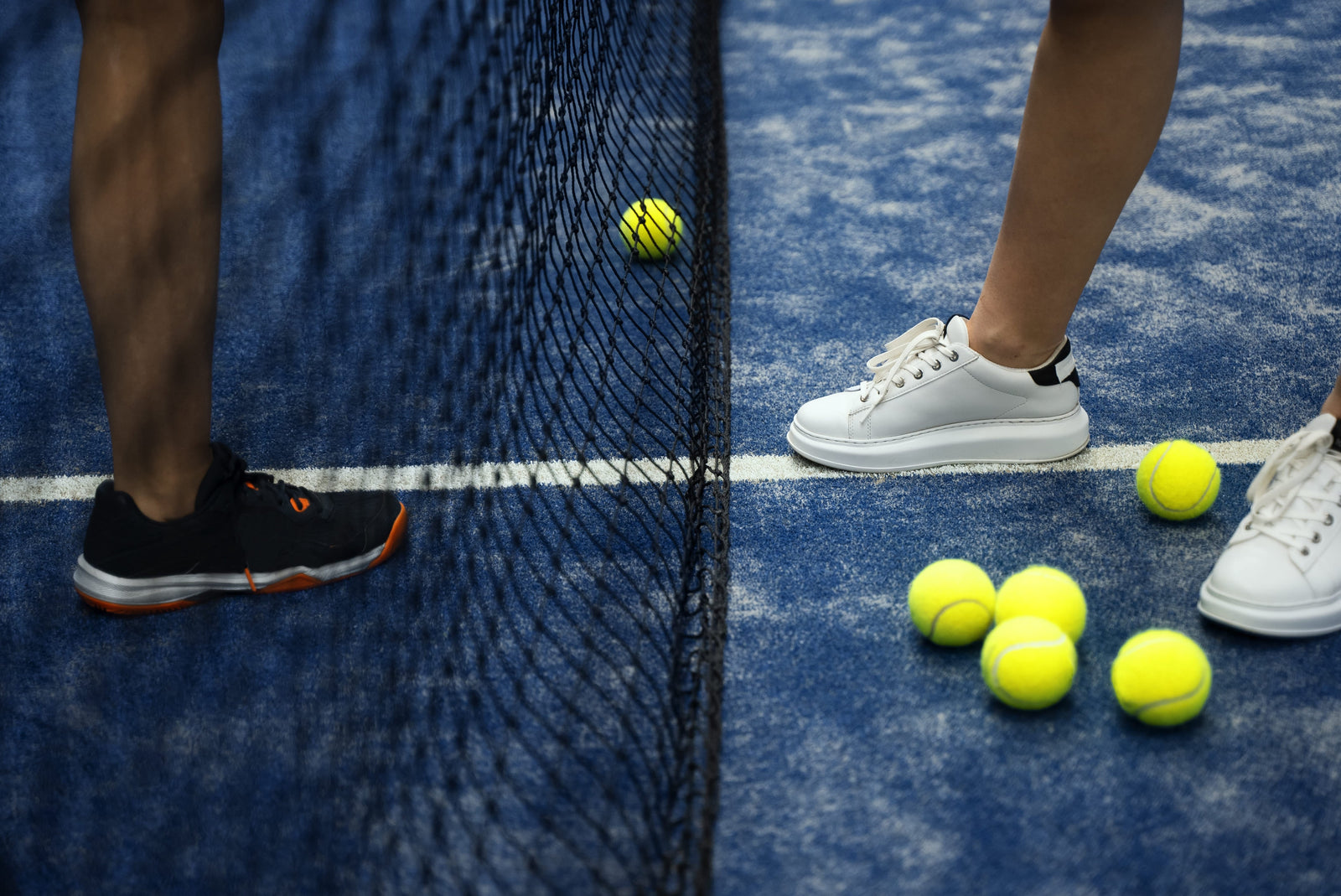 Close-up comparison of padel vs tennis shoes on a blue court with yellow balls and a net in between players.