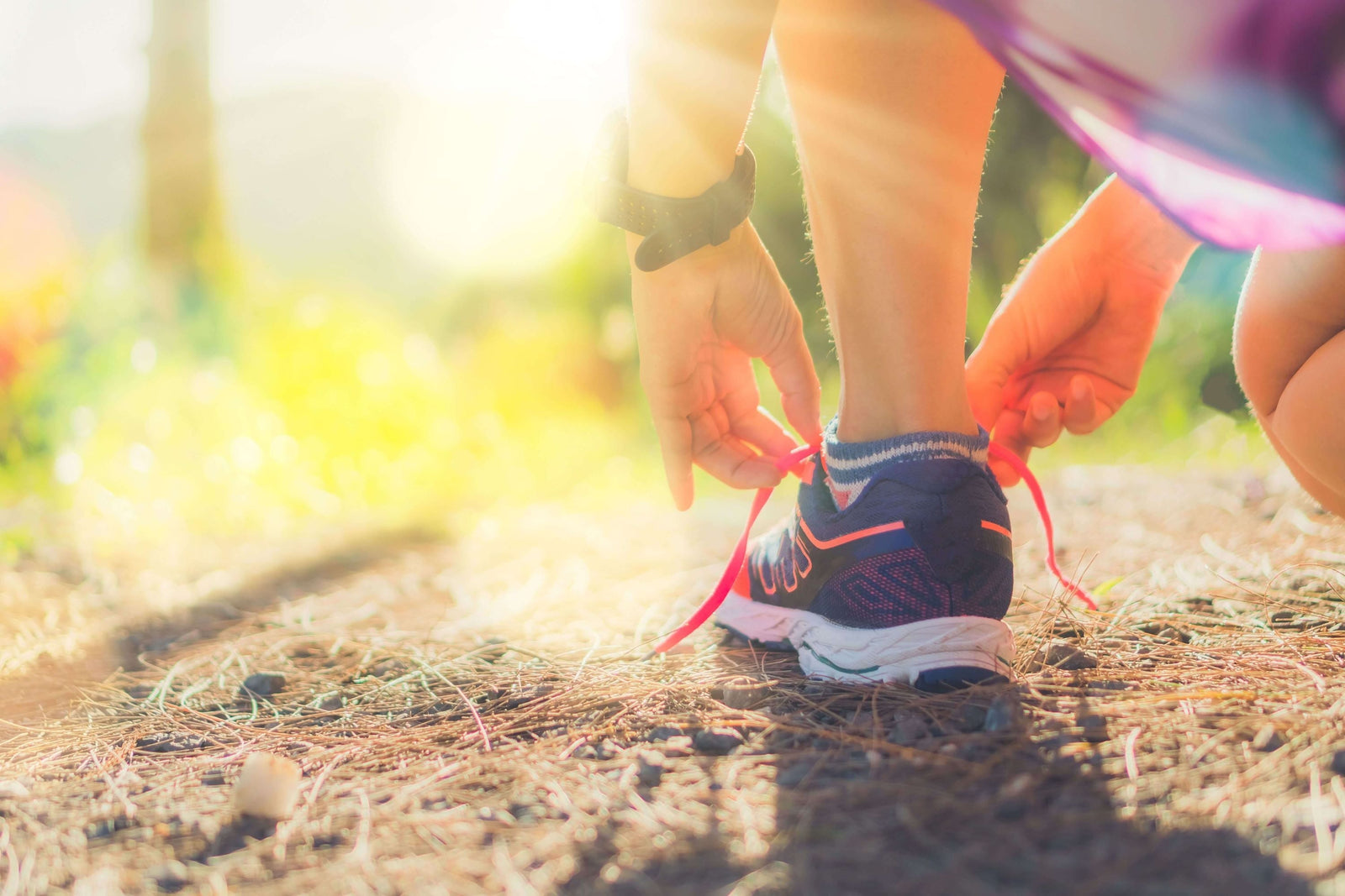 Lady in nature facing the sun and kneeling on the ground to lace up her navy and pink running shoe 